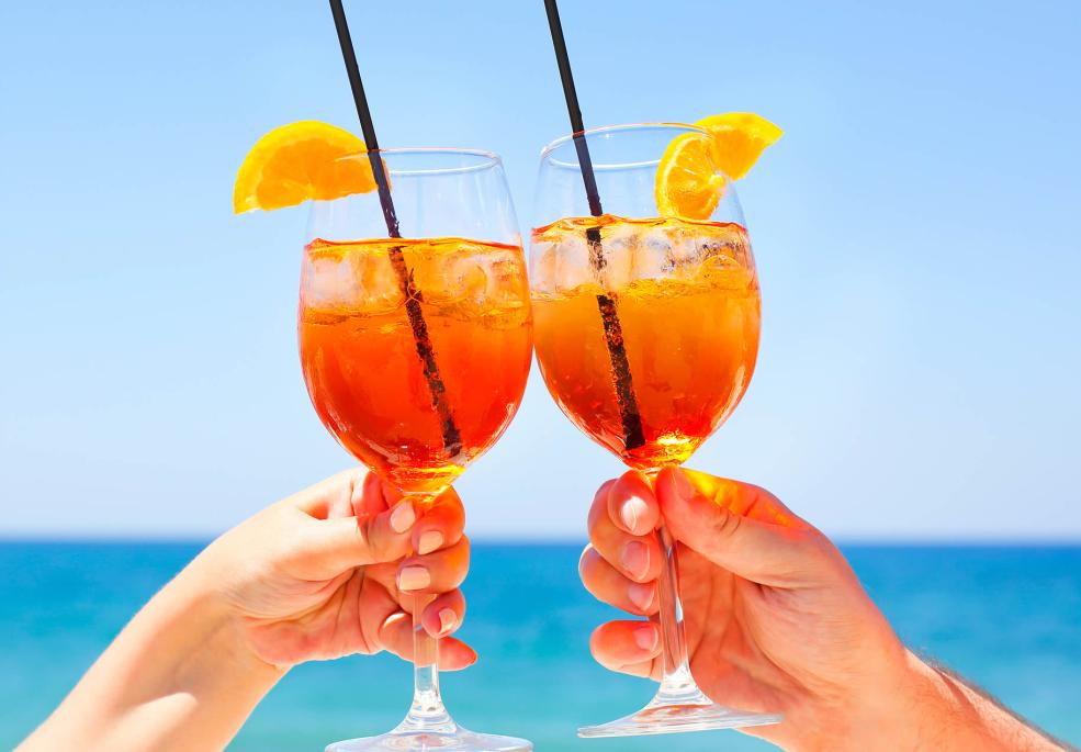 Due calici di spritz brindano al sole, con mare e cielo azzurro.